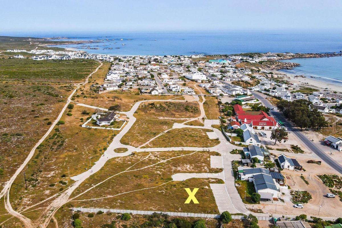 Aerial view of a vacant residential plot marked with an X, situated in a coastal development with established road infrastructure and nearby beach access.