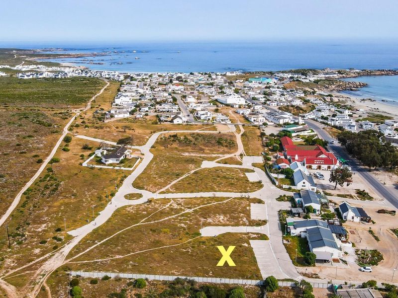 Aerial view of a vacant residential plot marked with an X, situated in a coastal development with established road infrastructure and nearby beach access.