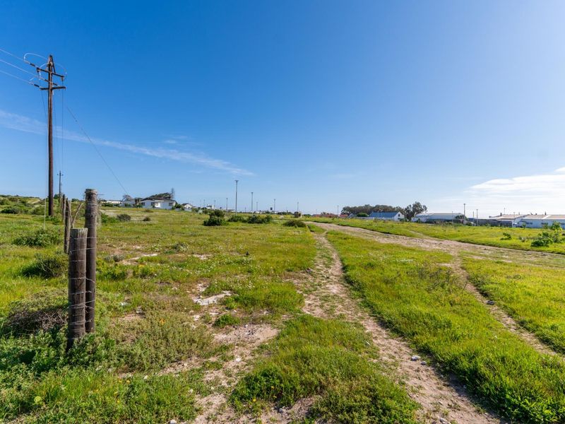A vacant residential plot featuring natural grass, dirt tracks, and existing utility infrastructure, offering a clear view of the surrounding neighborhood.