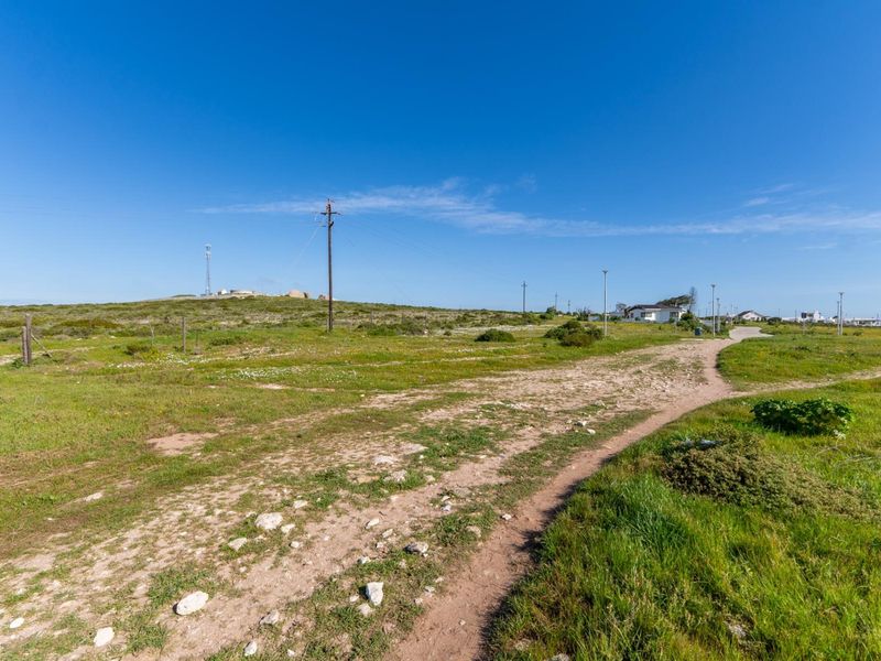A wide-angle view of a vacant residential plot featuring natural vegetation, a dirt access path, and existing utility infrastructure.