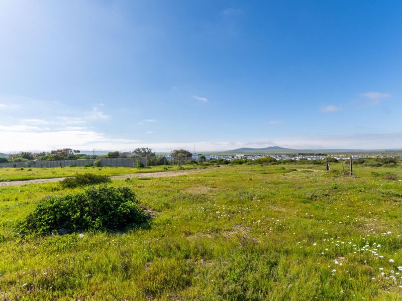 A wide-angle view of a vacant residential plot featuring natural vegetation, perimeter fencing, and distant landscape views under a clear blue sky.