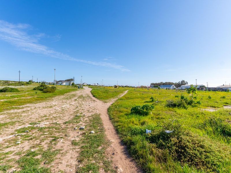 A view of a vacant residential plot featuring a dirt path, natural grassland, and surrounding neighborhood infrastructure.
