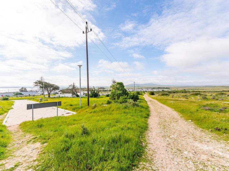 A view of a vacant residential plot featuring a gravel access path, paved parking area, and existing utility infrastructure against a natural landscape backdrop.