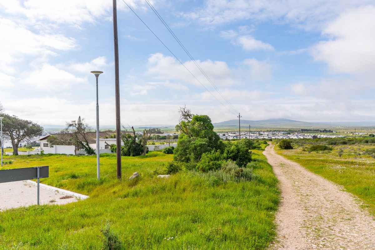 A view of an unpaved access road leading through a vacant residential plot with utility infrastructure and distant landscape views.