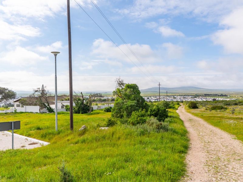 A view of an unpaved access road leading through a vacant residential plot with utility infrastructure and distant landscape views.