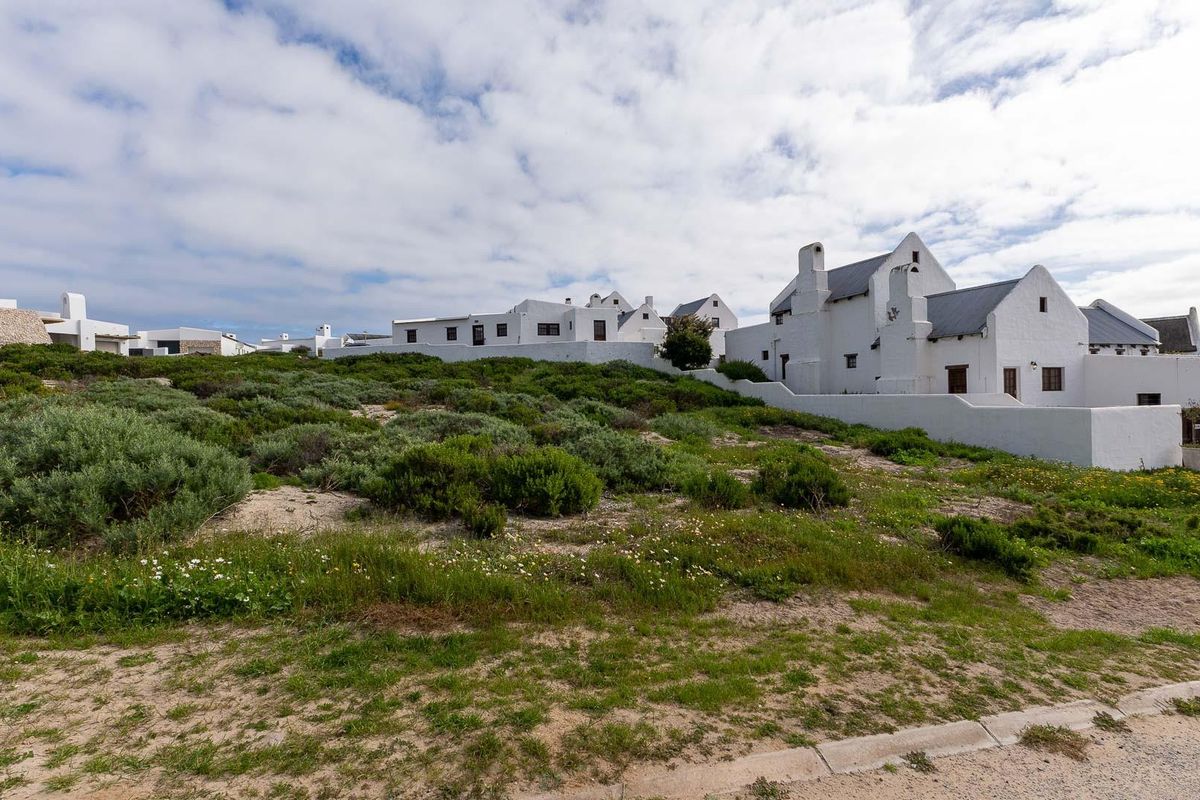 A view of a residential property featuring white-walled buildings and natural coastal vegetation, situated along a paved road.