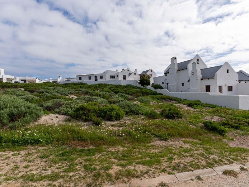 A view of a residential property featuring white-walled buildings and natural coastal vegetation, situated along a paved road.