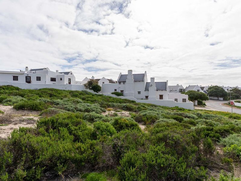 A view of a vacant residential plot featuring coastal vegetation and sand dunes, situated adjacent to an established residential housing development and paved road.
