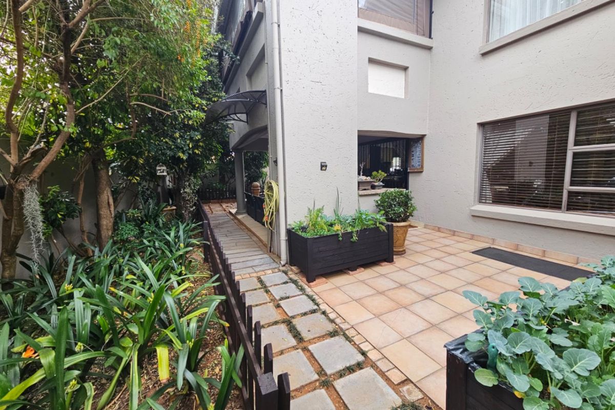 A well-maintained rear townhouse patio featuring tiled flooring, a paved walkway, and established garden landscaping with a timber fence.
