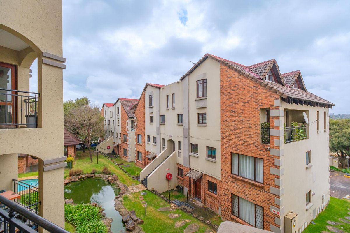 View of the apartment complex rear elevation featuring brick and plaster facade, tiled roofing, external staircases, and a landscaped garden with a pond.