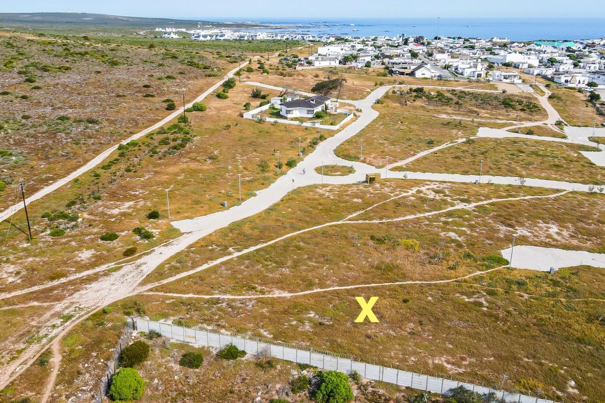 Aerial view of a vacant residential plot marked with an X, featuring surrounding paved roads, infrastructure, and proximity to the coastal town.