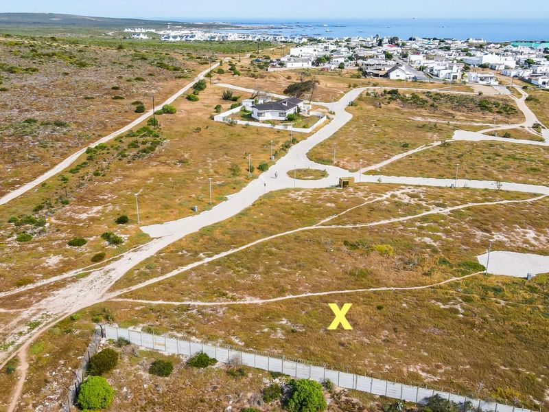 Aerial view of a vacant residential plot marked with an X, featuring surrounding paved roads, infrastructure, and proximity to the coastal town.