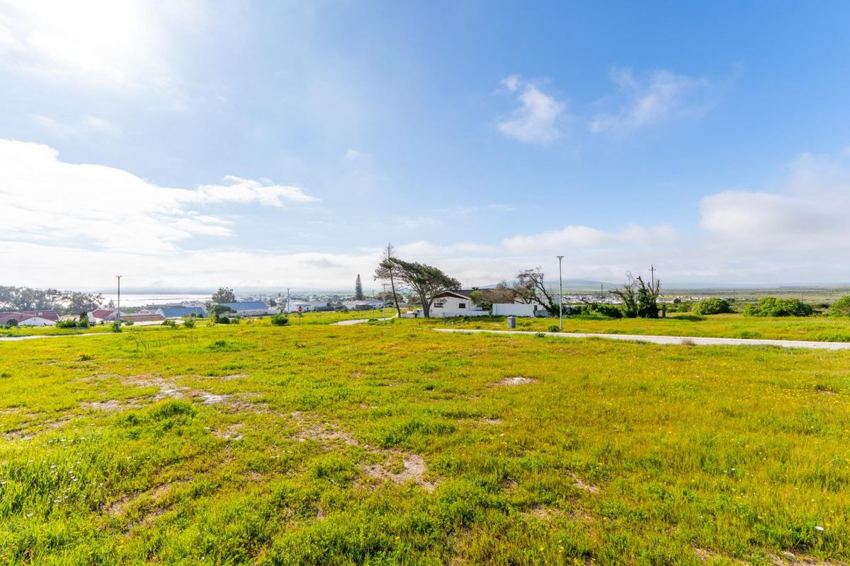 A wide-angle view of a vacant residential plot featuring natural grass coverage, paved road access, and distant coastal views.