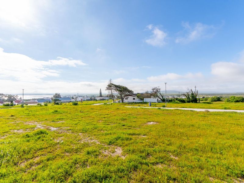 A wide-angle view of a vacant residential plot featuring natural grass coverage, paved road access, and distant coastal views.