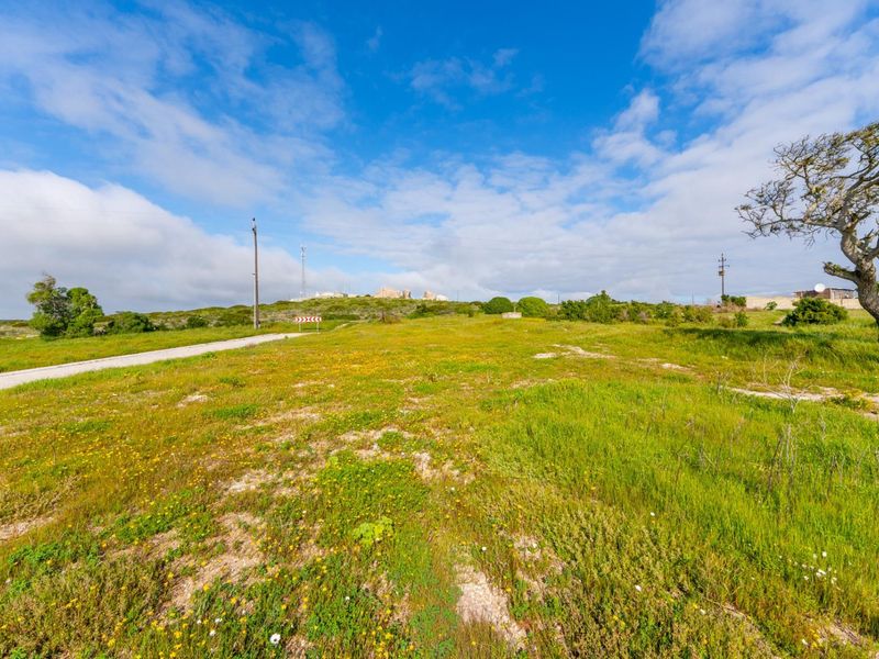 A vacant residential plot featuring natural vegetation, a gravel access road, and existing utility infrastructure, offering a clear site for future development.