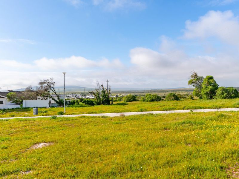 A wide-angle view of a vacant residential plot featuring natural grass, a paved road, and distant landscape views.