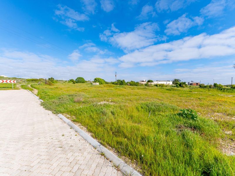 A vacant residential plot featuring a paved driveway access point, natural grass coverage, and surrounding neighborhood infrastructure under a clear blue sky.