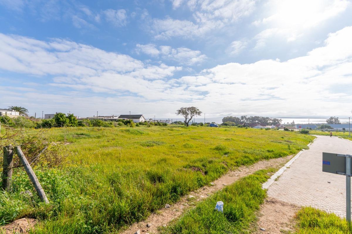 A vacant residential plot featuring natural grassland, bordering a paved sidewalk with distant coastal views.