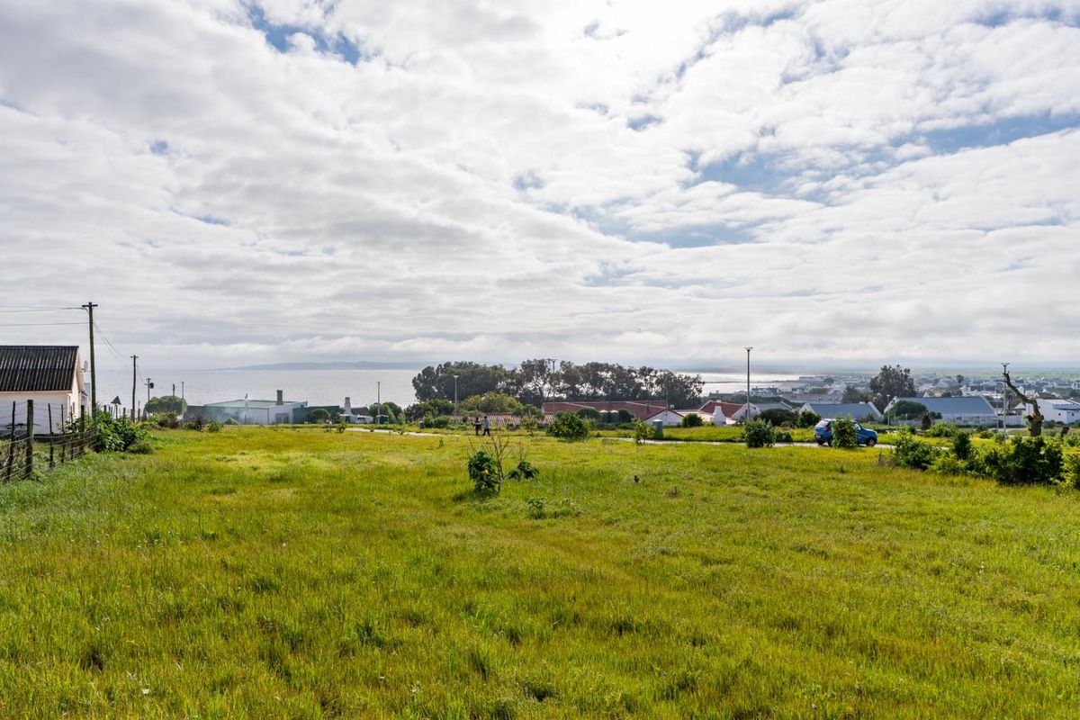 A wide-angle view of a vacant residential plot featuring grassy terrain, utility infrastructure, and distant coastal scenery.