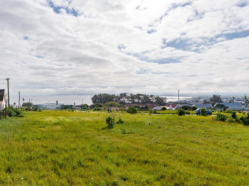 A wide-angle view of a vacant residential plot featuring grassy terrain, utility infrastructure, and distant coastal scenery.