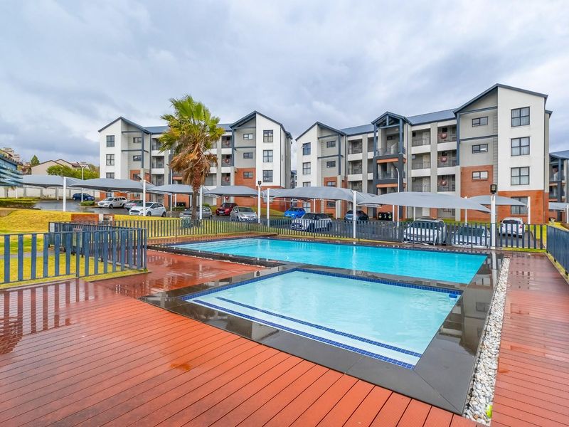 Communal swimming pool area featuring a wading pool, timber decking, and secure metal fencing, set within a modern apartment complex with covered parking.