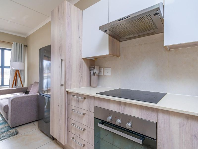 Modern open-plan kitchen featuring light wood-finish cabinetry, stone countertops, integrated oven and hob, flowing into a bright living area with large windows.