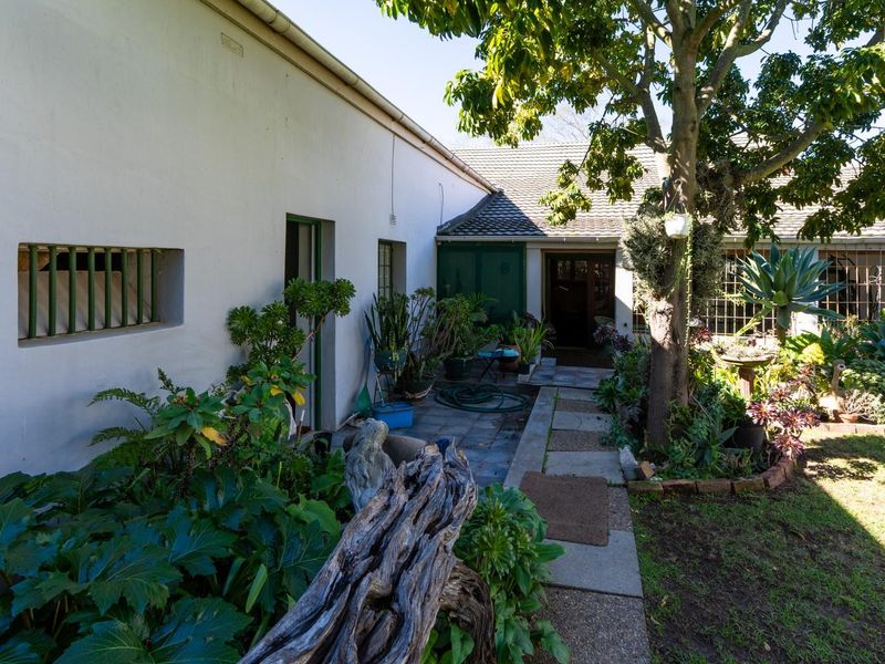 A rear garden view featuring a paved walkway leading to a covered patio area with established landscaping and mature trees.