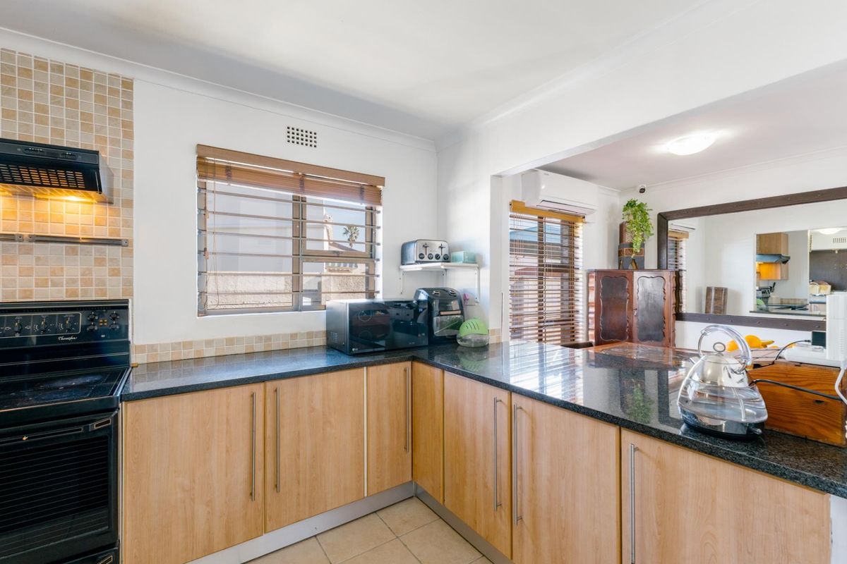 A functional kitchen featuring light wood-tone cabinetry, dark granite countertops, a built-in oven with extractor fan, and tiled splashbacks, opening into an adjacent living area.