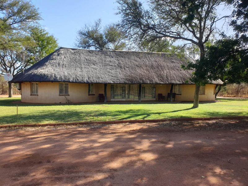 A traditional farmhouse featuring a classic thatched roof, a covered veranda, and large sliding glass doors set within a spacious, tree-lined garden.