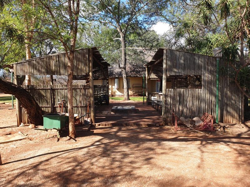 Outdoor entertainment area featuring a rustic thatched-roof structure, brick paving, and a central built-in fire pit, set within a natural farm landscape.
