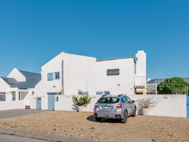 Front exterior view of a white-walled house featuring a gravel driveway, boundary wall, and blue shuttered windows under a clear blue sky.
