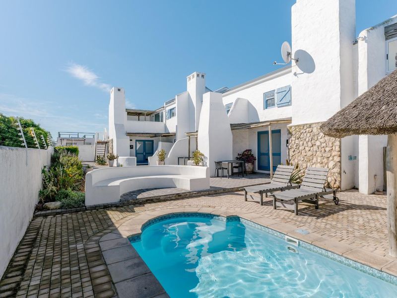 A well-maintained outdoor pool area featuring a paved patio, built-in curved seating, stone feature wall, and a thatched umbrella.