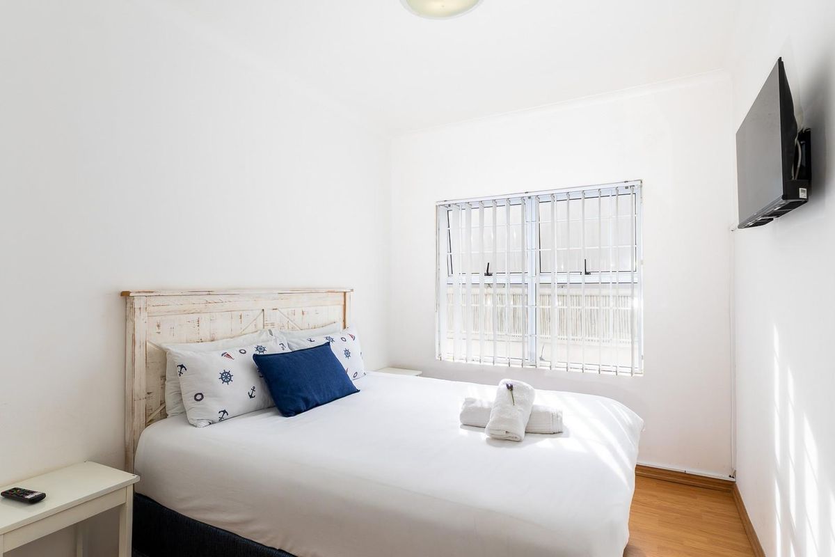 A bright bedroom featuring laminate flooring, vertical window blinds with security bars, and a wall-mounted television.