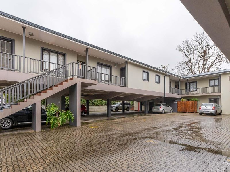 Exterior view of an apartment complex featuring a paved parking area, covered parking bays, and an external staircase leading to upper-level units.