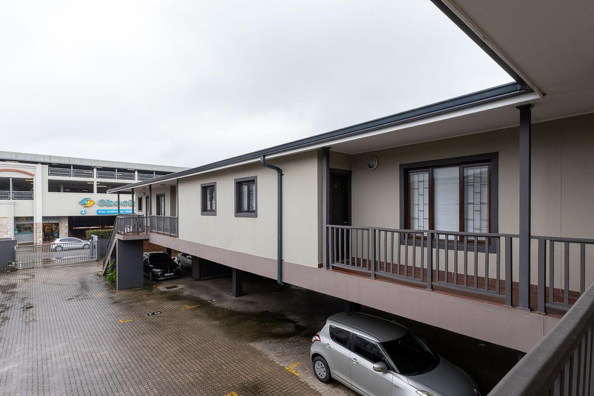 Exterior view of an apartment complex featuring a covered walkway, metal railings, and a paved ground-level parking area.