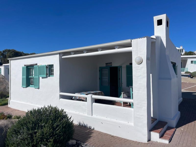 A charming white cottage featuring a covered front patio, traditional shutters, and a prominent chimney, set against a clear blue sky.
