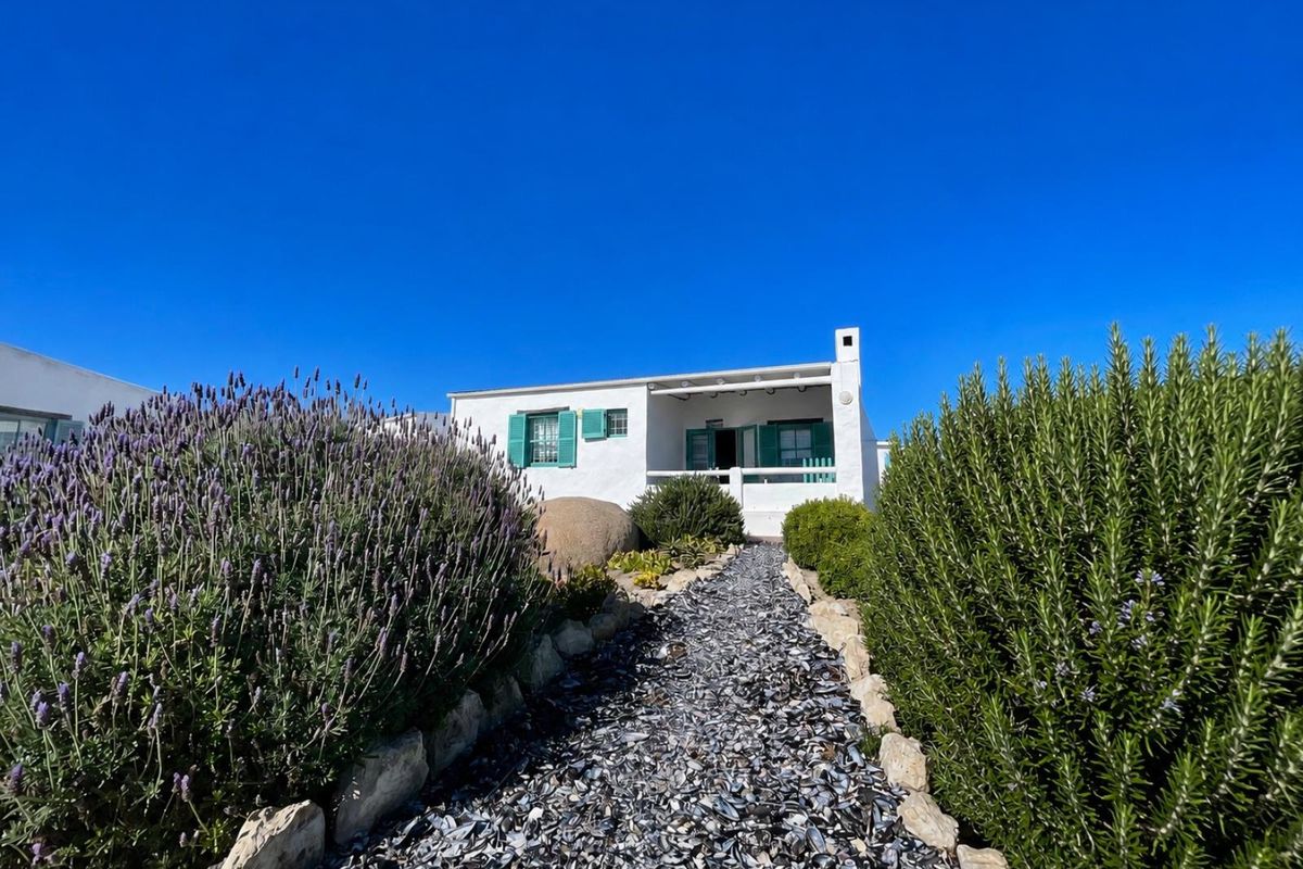 A charming white cottage exterior featuring green shutters, a covered patio, and a unique shell-lined pathway leading through a landscaped garden.