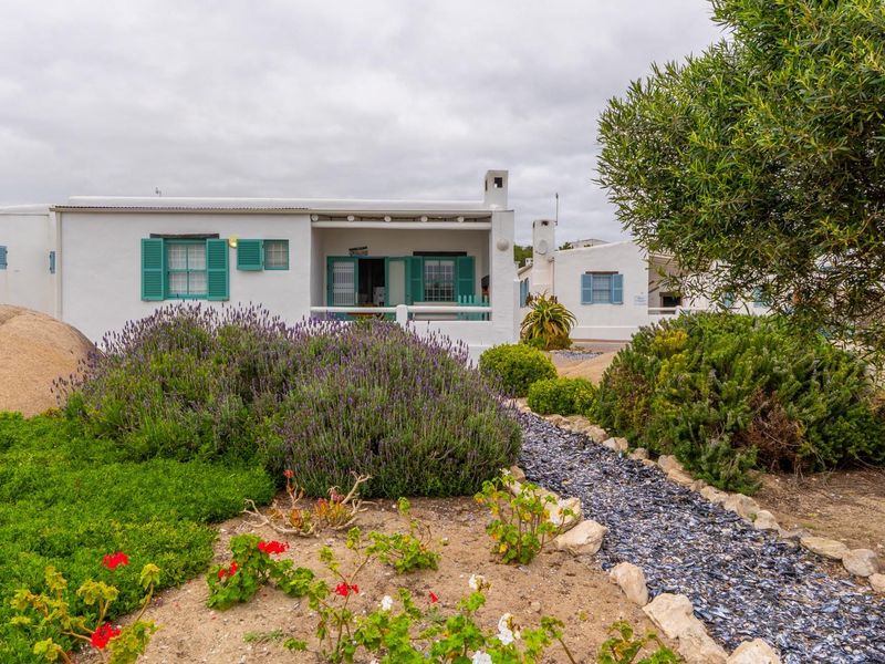 A charming rear exterior view of a white cottage featuring a covered patio, traditional green shutters, and a well-maintained landscaped garden with stone pathways.
