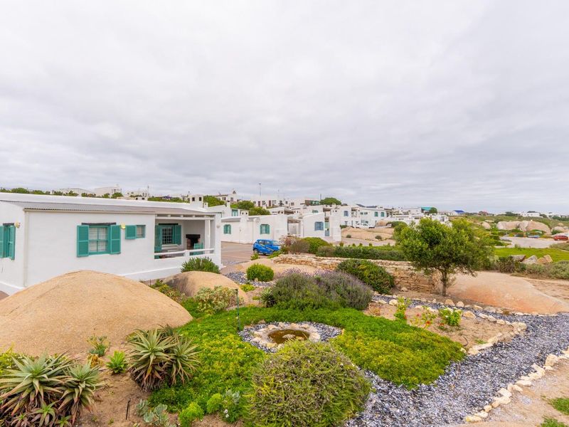 A well-maintained coastal cottage featuring a covered patio, charming green shutters, and a landscaped garden with natural rock formations in a scenic estate setting.