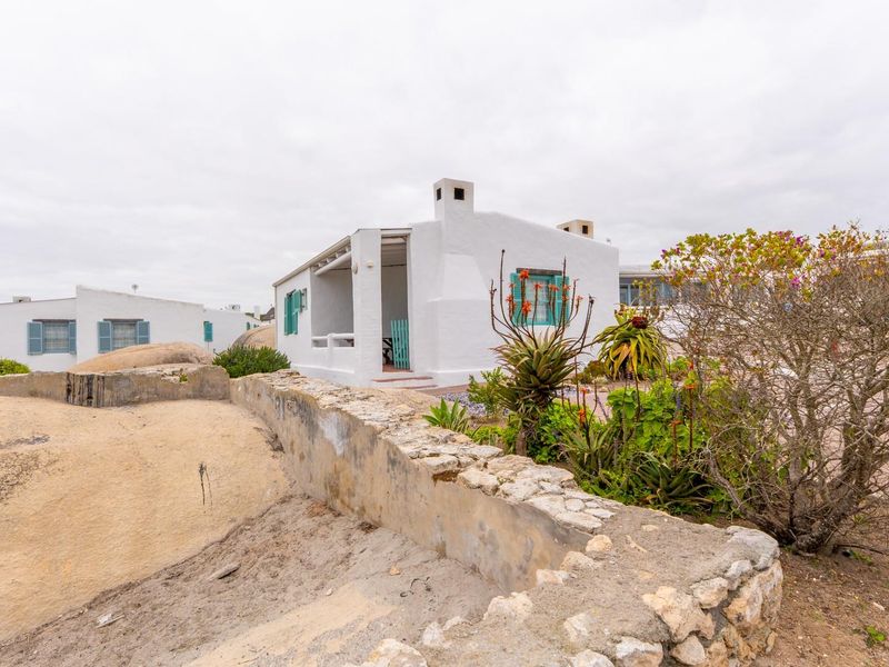 A view of the rear exterior of a white-walled coastal cottage featuring a stone retaining wall and a covered patio area.