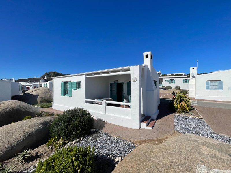 A charming white cottage featuring a covered front patio, traditional shutters, and a chimney, set within a well-maintained coastal estate environment.