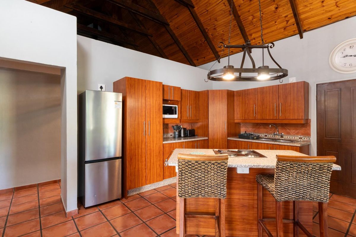 A kitchen featuring wooden cabinetry, granite countertops, a central island with a built-in hob, and a vaulted timber ceiling with terracotta flooring.