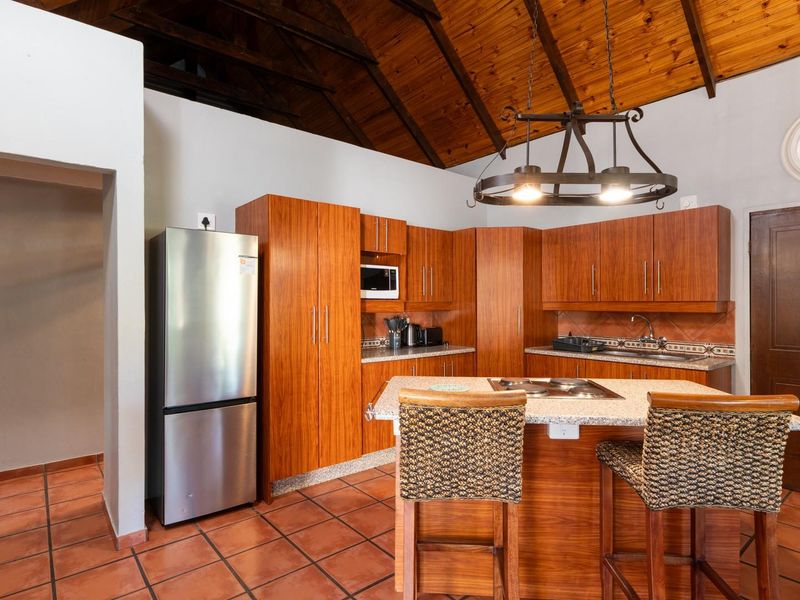 A kitchen featuring wooden cabinetry, granite countertops, a central island with a built-in hob, and a vaulted timber ceiling with terracotta flooring.