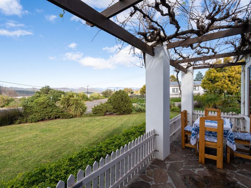 A charming covered patio featuring a timber pergola, stone flooring, and a picket fence, overlooking a well-maintained garden and scenic mountain views.