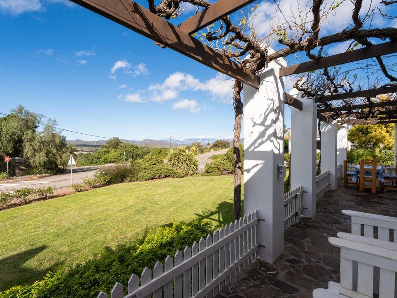A covered patio featuring a timber pergola, stone flooring, and a picket fence, overlooking a landscaped garden and scenic mountain views.