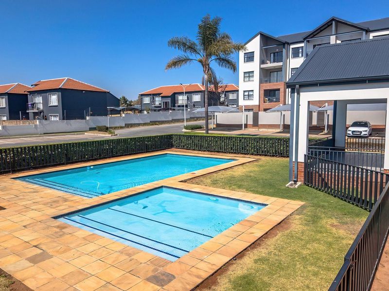 A well-maintained communal swimming pool area featuring paved decking, metal fencing, and an adjacent covered patio within a modern apartment complex.