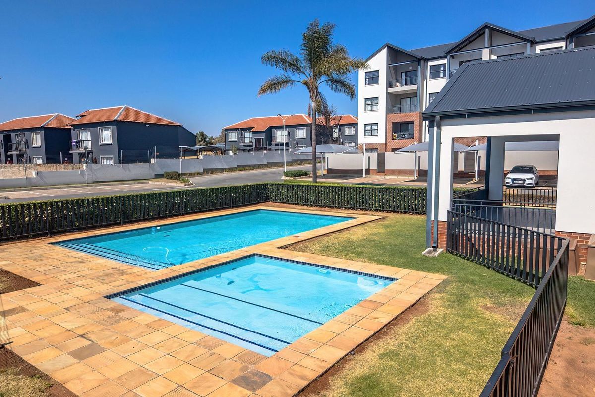 A well-maintained communal swimming pool area featuring paved decking, metal fencing, and an adjacent covered patio within a modern apartment complex.