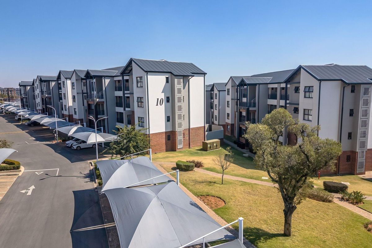 Aerial view of a modern apartment complex featuring multi-story residential buildings, paved parking areas with shade cloth carports, and well-maintained communal garden spaces.