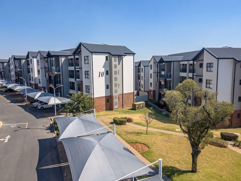 Aerial view of a modern apartment complex featuring multi-story residential buildings, paved parking areas with shade cloth carports, and well-maintained communal garden spaces.