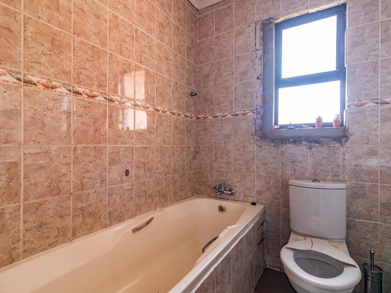 A dated bathroom featuring a built-in bathtub and toilet, with ceramic wall tiling and a window requiring minor plaster repairs.